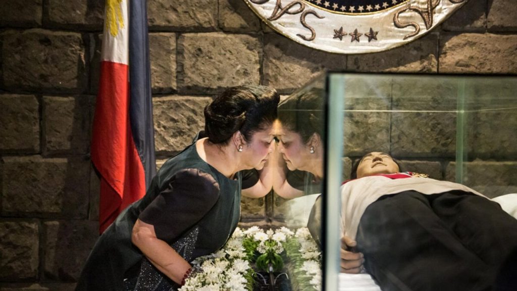 Imelda at her dead husband's glass coffin