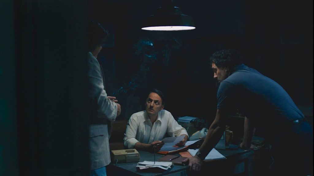 Ivan in a dark room with two heavies