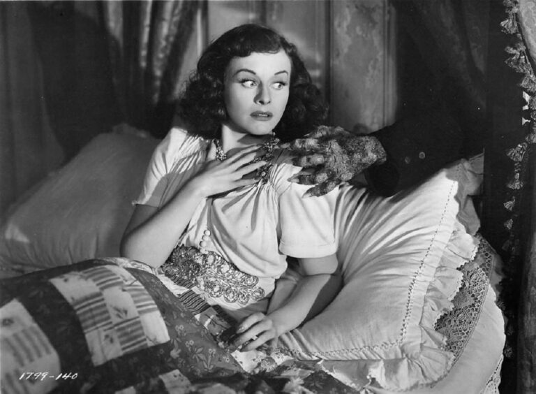 Joyce in bed with a disembodied hand making for her throat